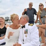 A newly married couple shares a kiss aboard a sailboat while guests raise drinks around them during a celebration on the Detroit River.