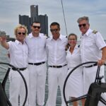 Five Detroit Charter Company crew members in white uniforms pose together at the helm of a sailboat with the Detroit skyline in the background.