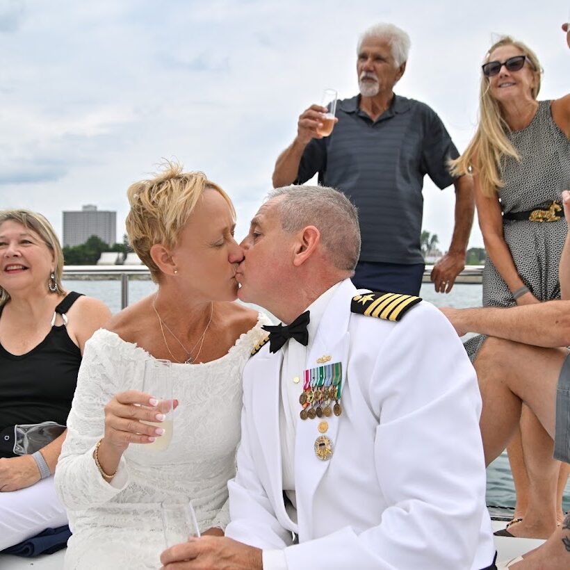 A newly married couple shares a kiss aboard a sailboat while guests raise drinks around them during a celebration on the Detroit River.