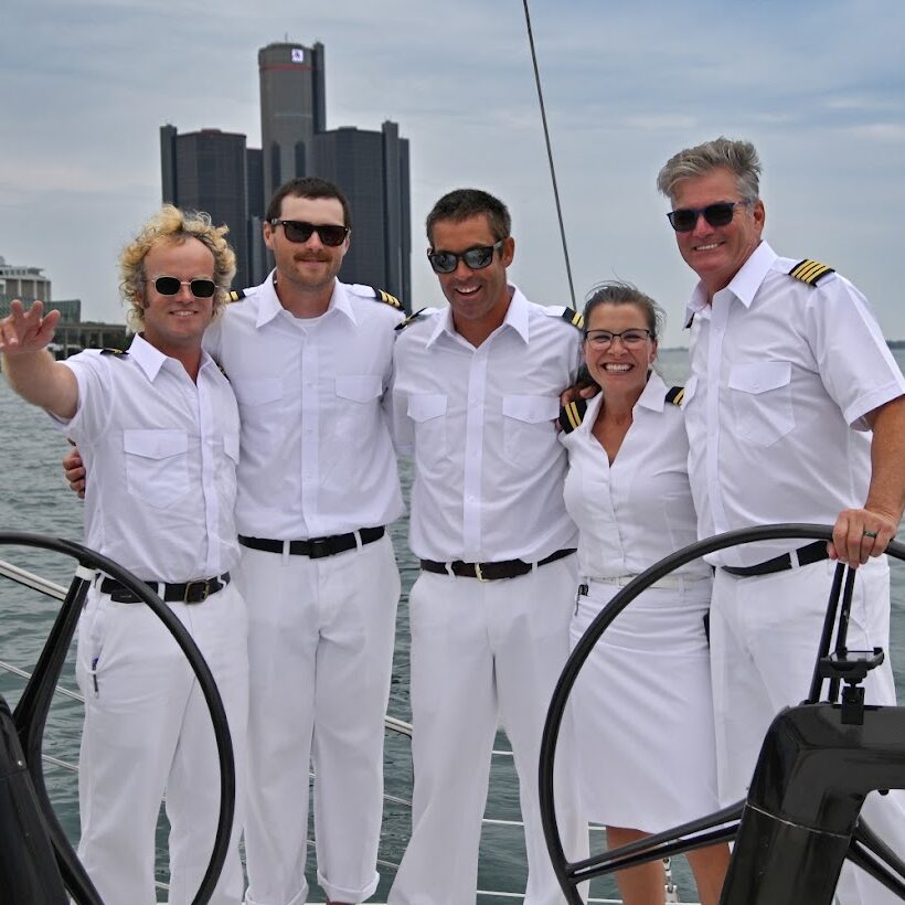 Five Detroit Charter Company crew members in white uniforms pose together at the helm of a sailboat with the Detroit skyline in the background.